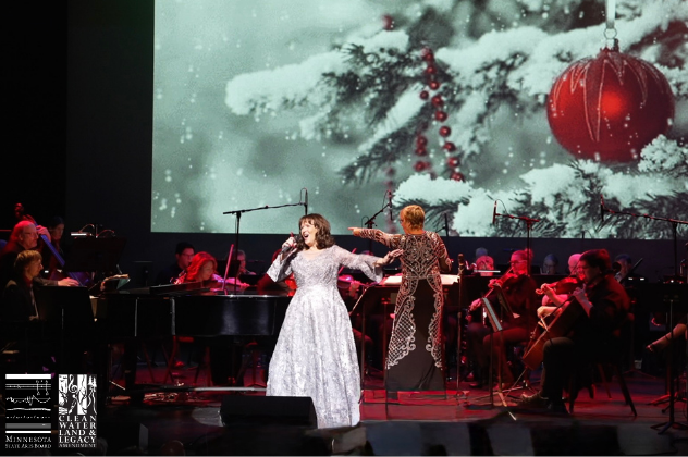 Colleen Raye A Holiday in Your Heart - Dec 8 woman singing on stage in front of a christmas scene and an orchestra